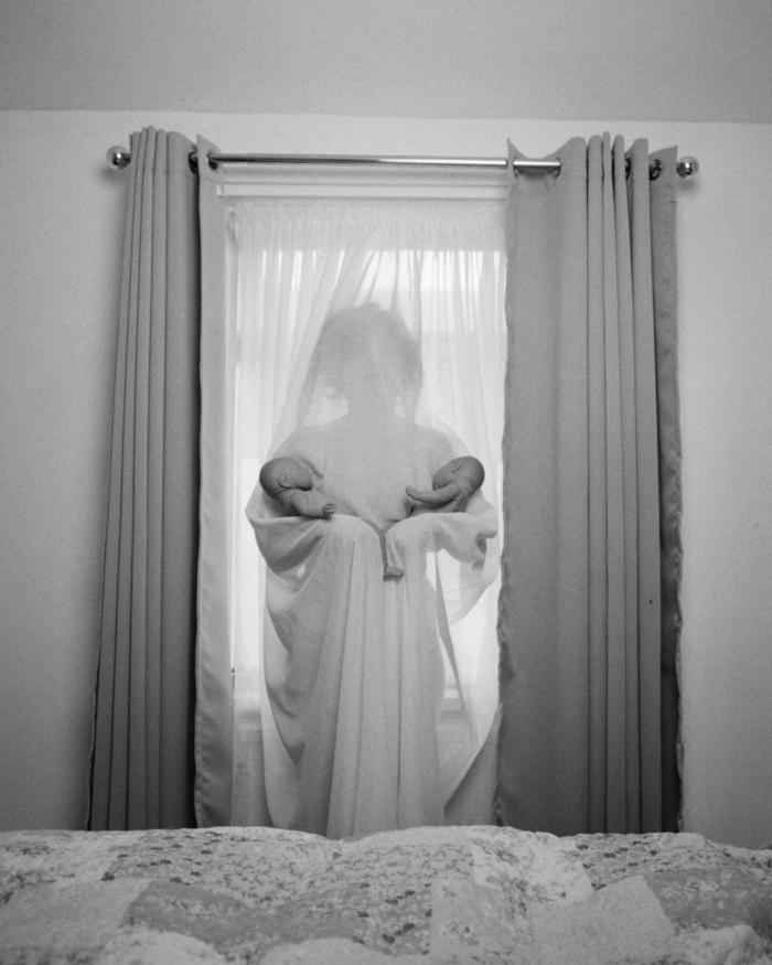 Bild: Amak MahmoodianEinhundertzwanzig Minuten, 2019–2024. Schwarz-Weiß-Fotografie einer Frau hinter einem Netzvorhang, die in jedem Arm ein Baby hält.